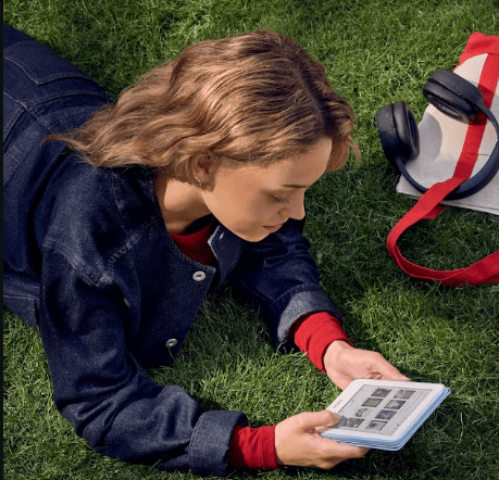 Person lying on grass, wearing a denim jacket, using a tablet with a red backpack beside them.
