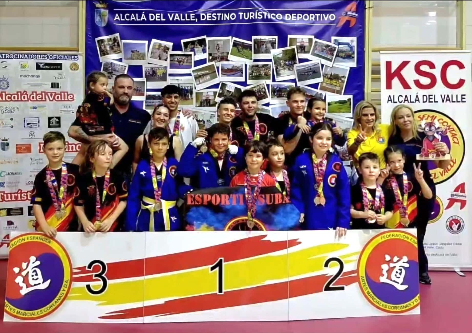 Group of young martial artists on a podium posing with medals after a competition in Alcalá del Valle, with banners and sponsors in the background.