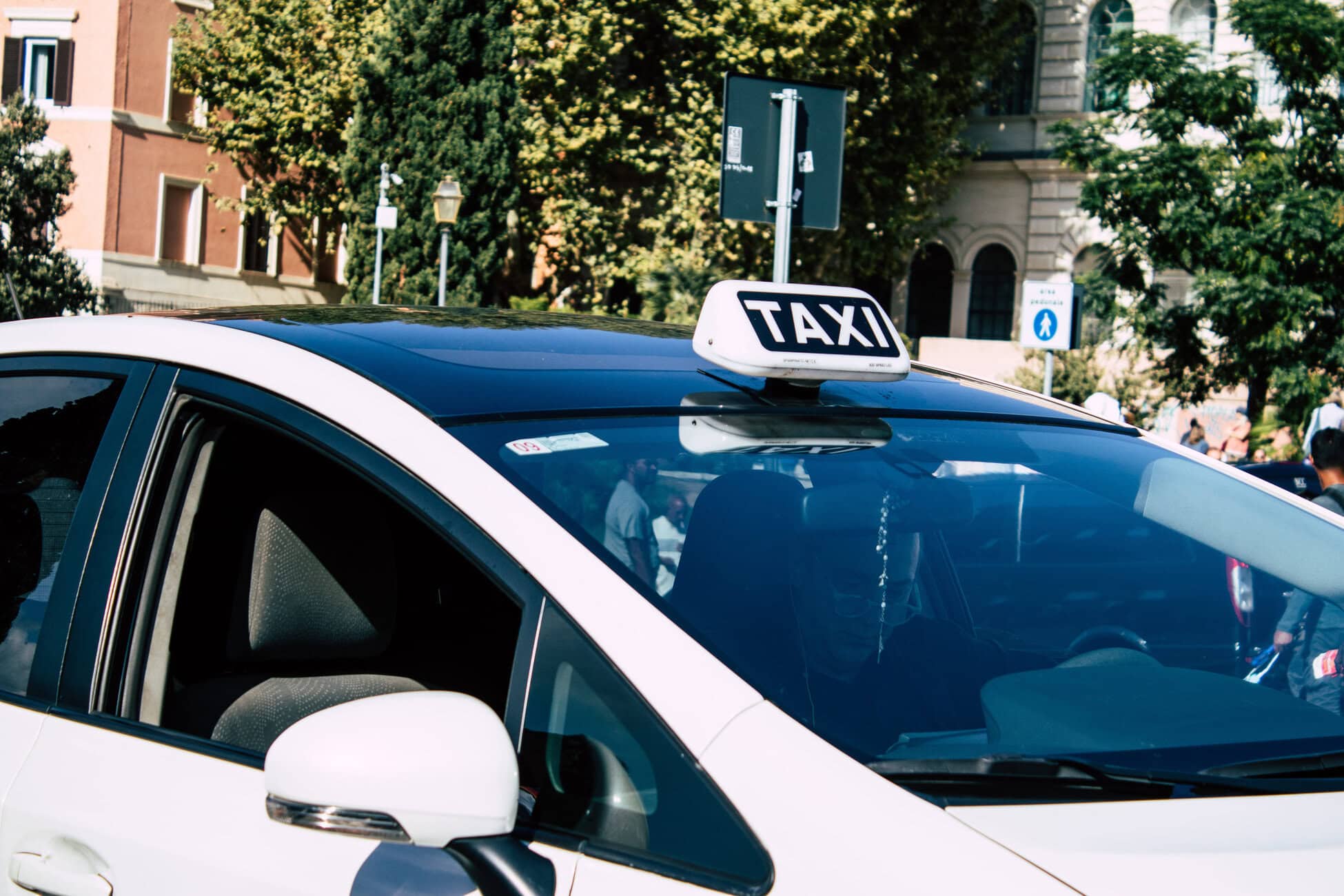 White taxi with a rooftop TAXI sign on a city street, with trees and brick buildings in the background.