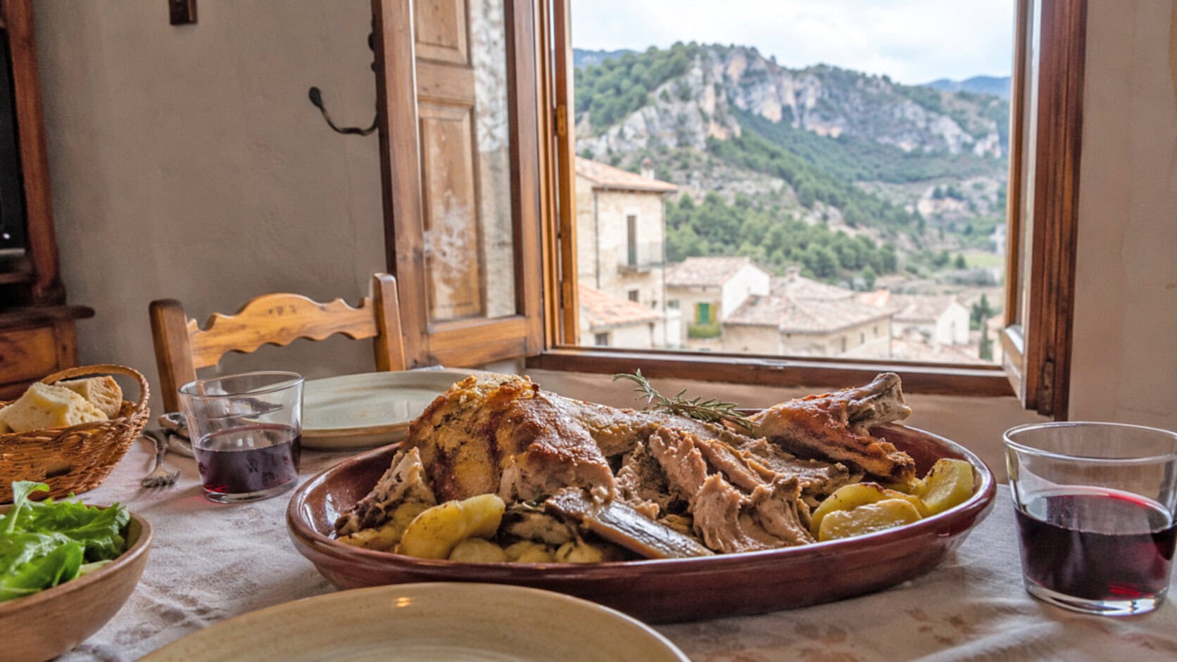 Table set with a roast meat, potatoes and rosemary, bread, and greens, by a window overlooking a hillside village.