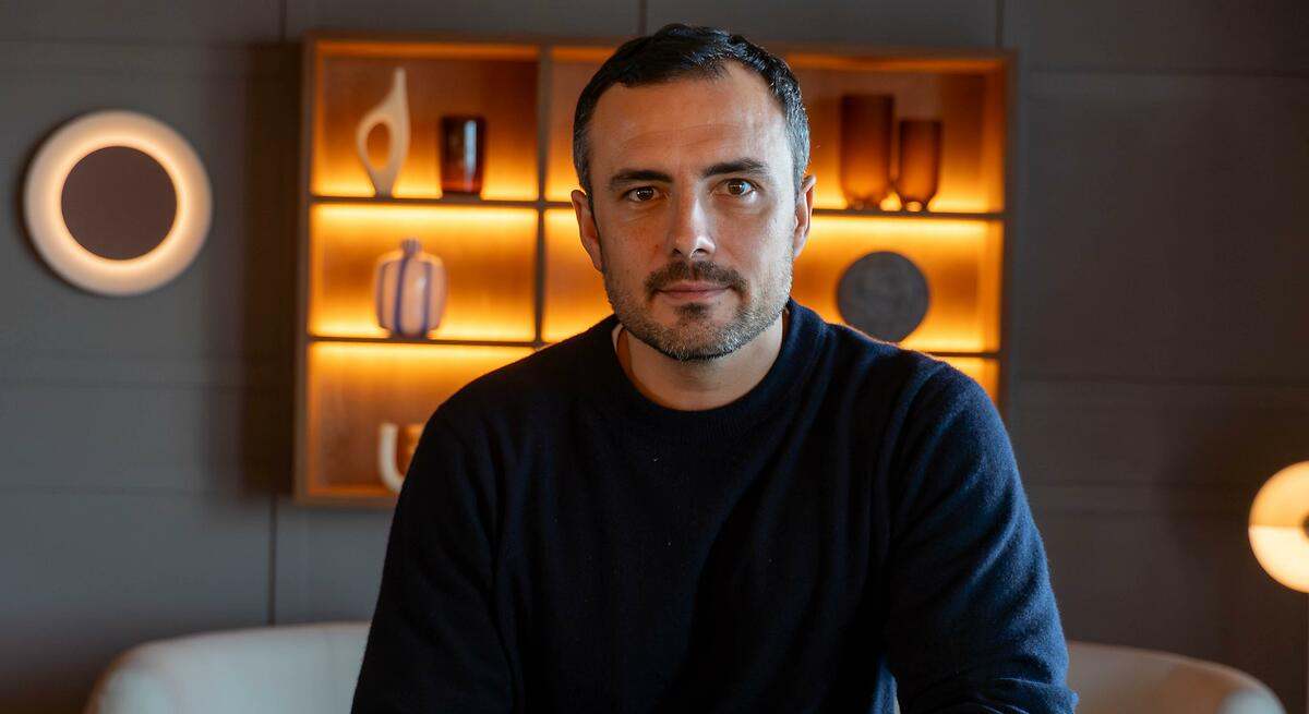 Portrait of a man with short dark hair and stubble sitting in a modern room, warm illuminated shelves behind him.