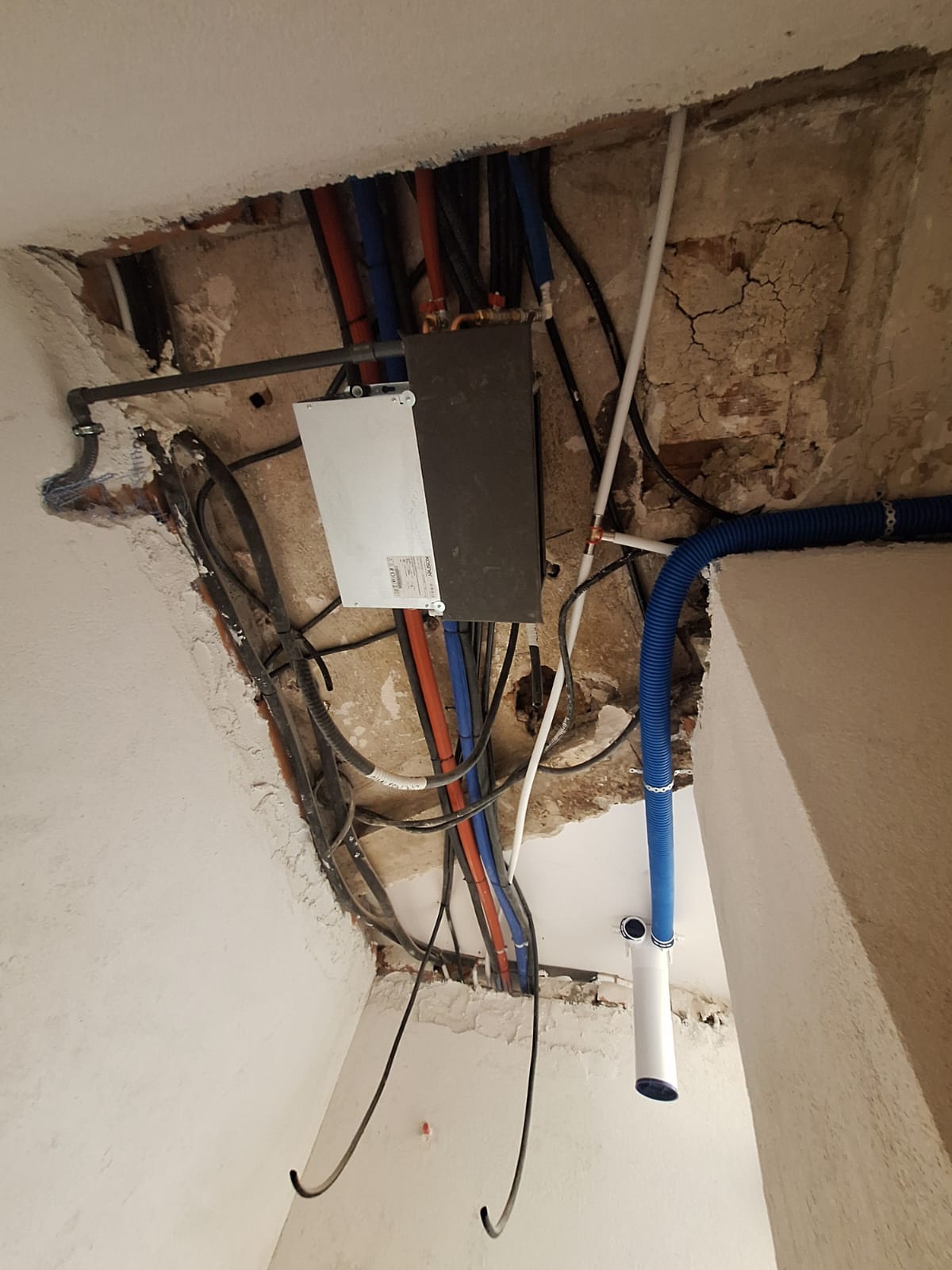 Open wall cavity with an electrical junction box and many tangled cables (blue, red, black) plus exposed plumbing pipes and a blue corrugated conduit nearby, in a damaged wall.
