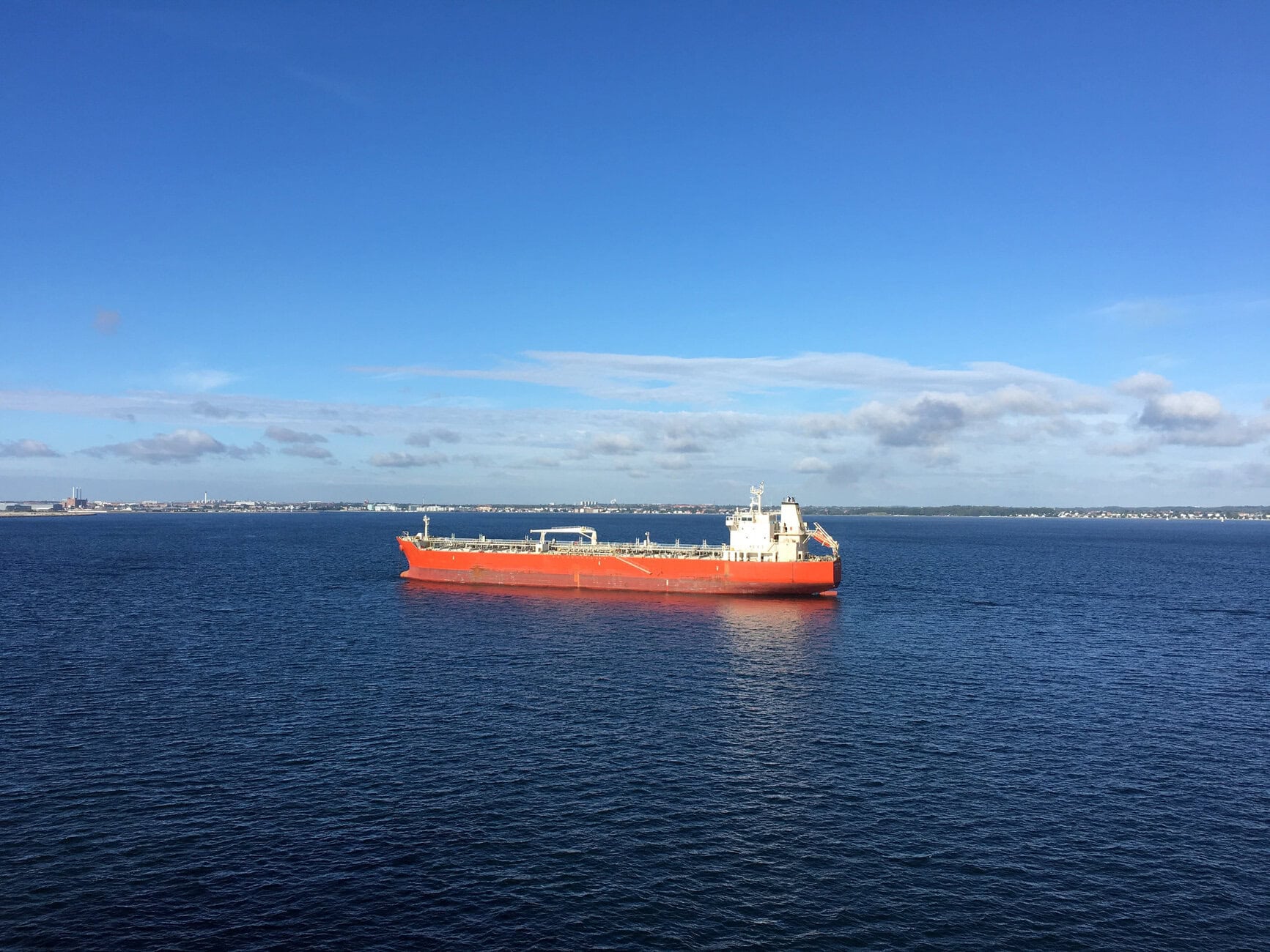 Orange cargo ship sailing on calm blue water with a distant city skyline and scattered clouds in the sky.