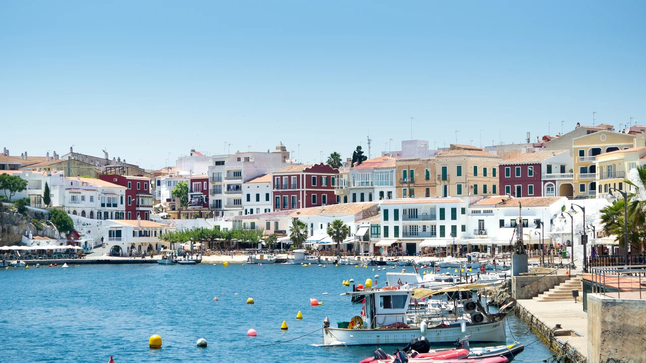 Colorful coastal town harbor with pastel buildings along the shore, boats docked at a calm blue sea under a clear sky.