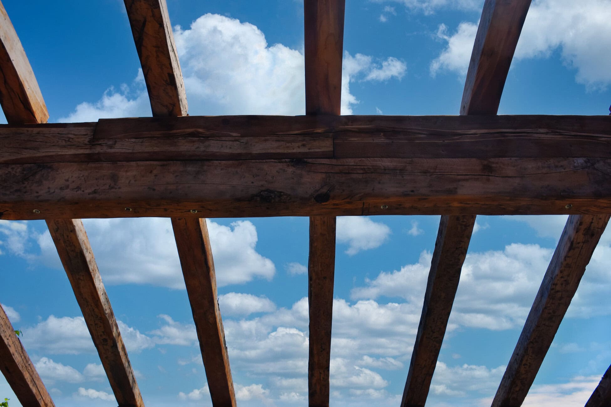 Wooden beam lattice framing a bright blue sky with white clouds, seen from below.