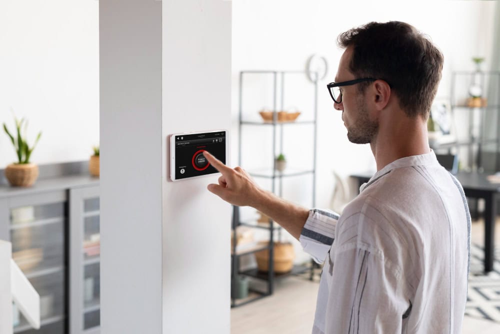 Man in glasses touches a wall-mounted smart home touchscreen on a white pillar in a bright living space.
