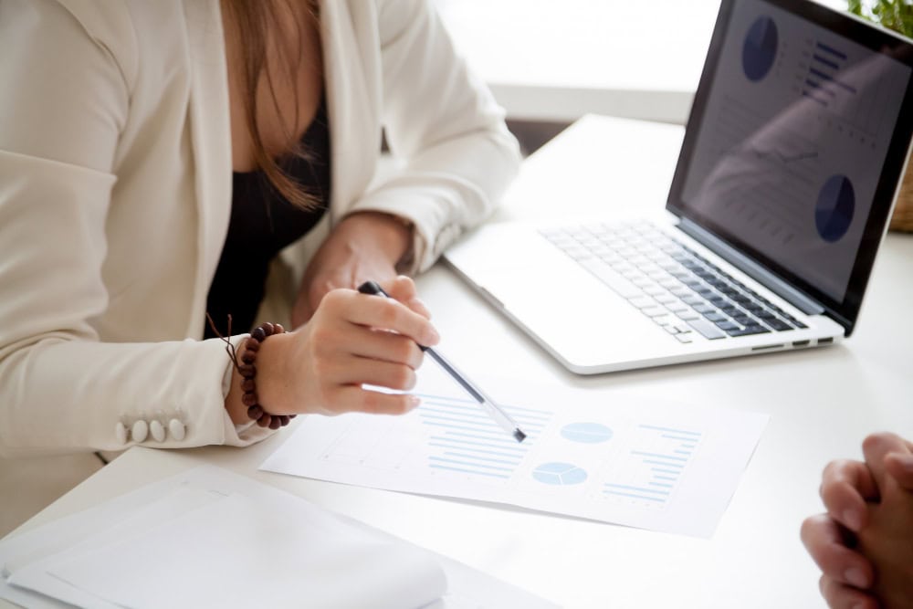 Person in a white blazer points at printed charts on a desk beside an open laptop, reviewing data.