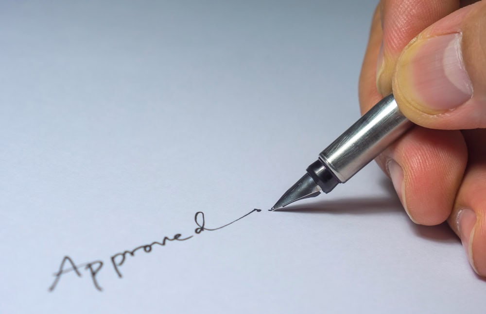 Close-up of a hand writing the word 'Approved' with a silver fountain pen on white paper.