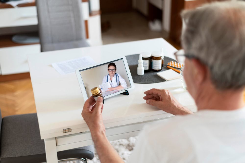 Senior person at a white table has a video call with a doctor on a tablet, holding a pill bottle as medicine bottles sit nearby at home