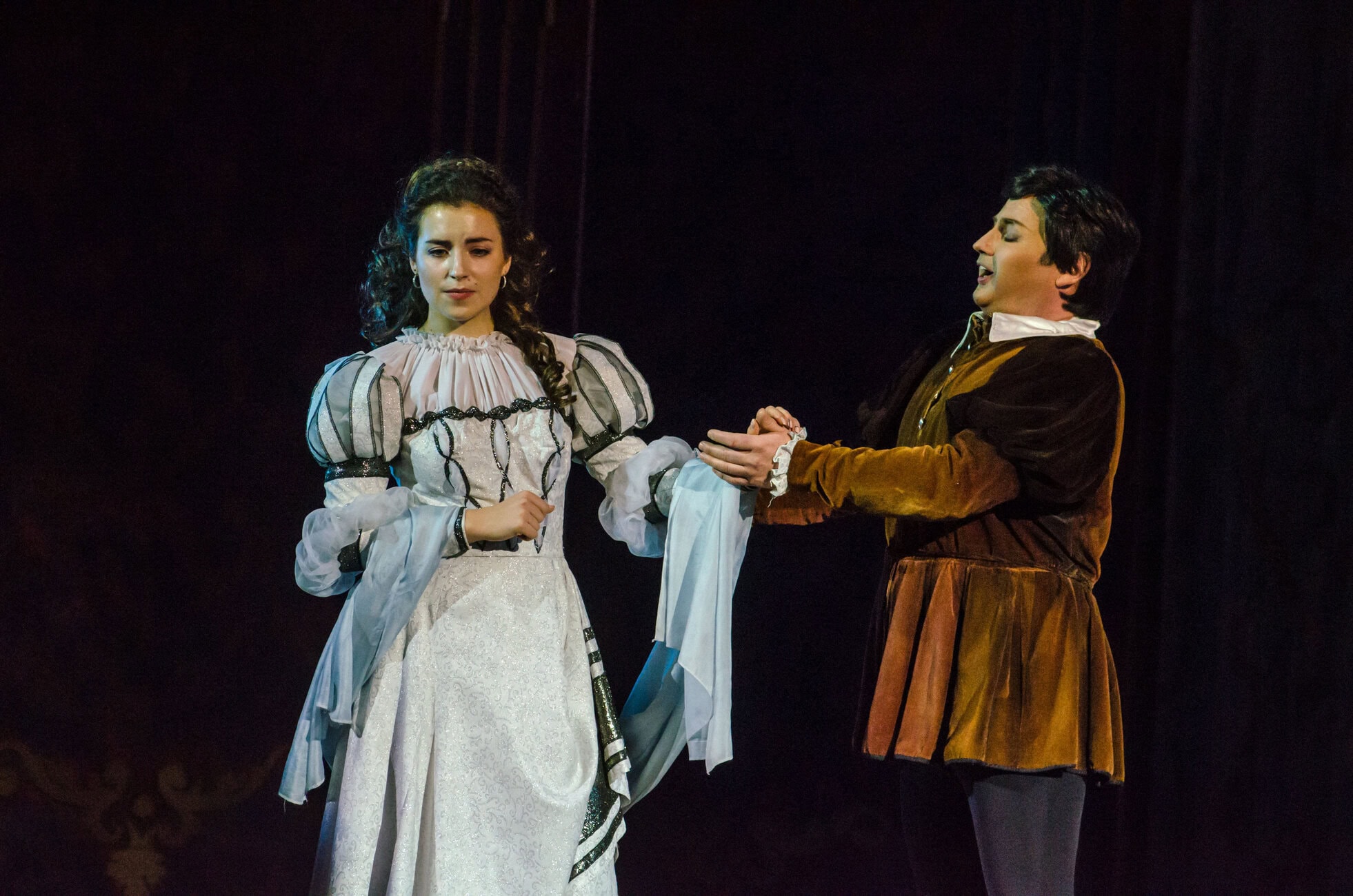 Two performers in period costumes hold hands on a dark stage: a woman in a white, ornate gown with puffy sleeves and braided hair, and a man in a brown historical outfit singing or speaking.