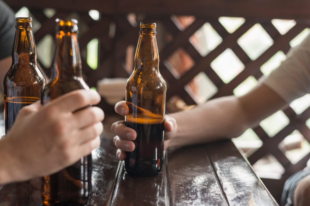 Three brown beer bottles on a wooden table as two people toast outdoors, hands holding bottles in a casual gathering.