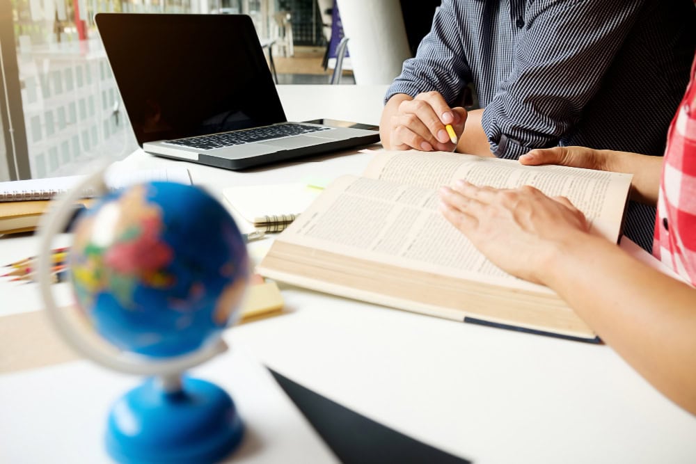 Two students study at a bright desk with a laptop, an open textbook, and a globe in the foreground.