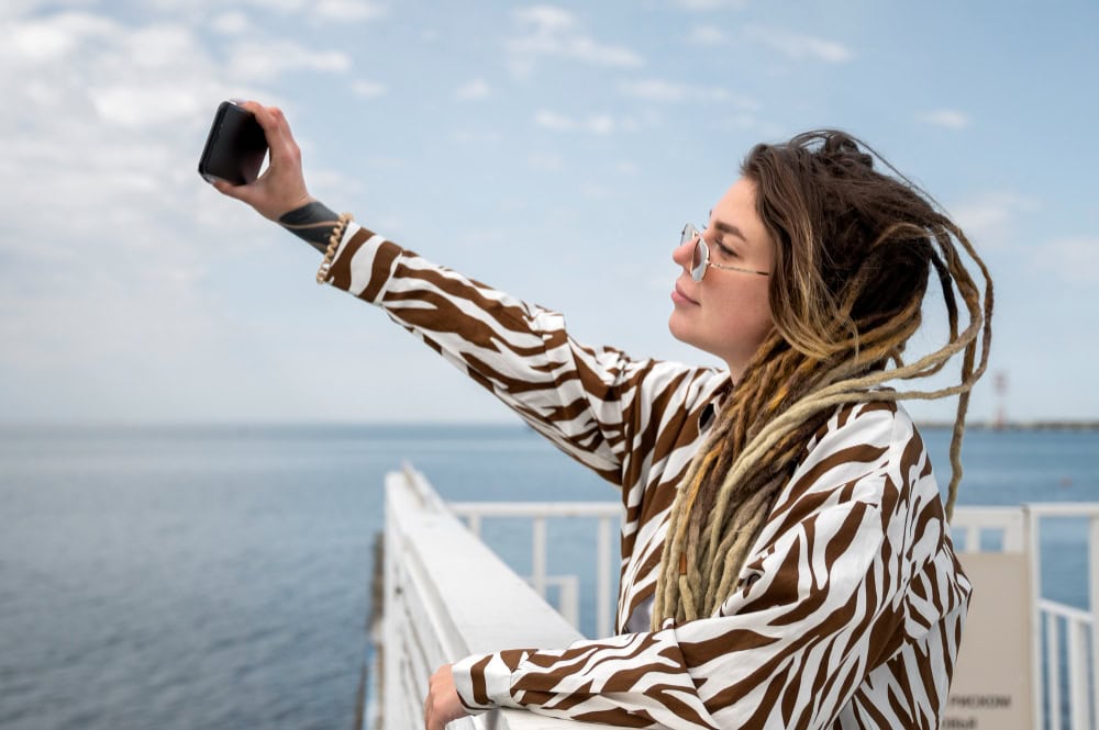 Woman with dreadlocks in a zebra-print shirt taking a selfie on a pier by the sea, wearing sunglasses.