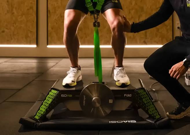 Person stands on a compact leg training device with a green strap securing the legs, trainer nearby providing support.