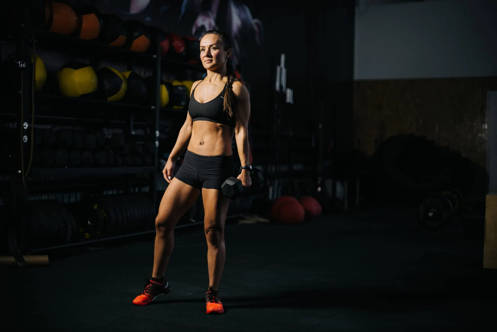 Fit woman in a black sports bra and shorts stands in a dimly lit gym, holding a dumbbell in one hand.