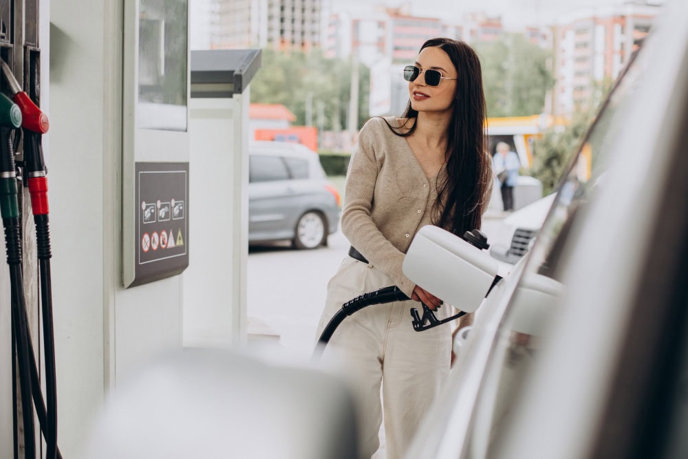 Woman wearing sunglasses and a beige cardigan refueling a white car at a gas pump at a station.