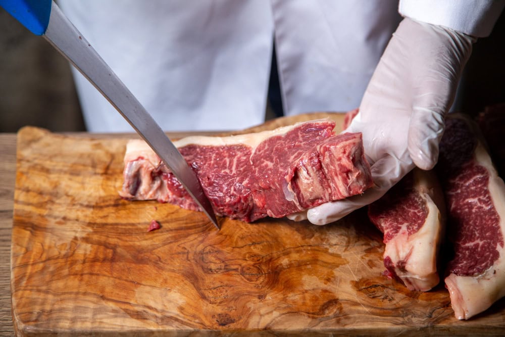 Butcher wearing gloves slicing a raw beef steak on a wooden cutting board with a large knife