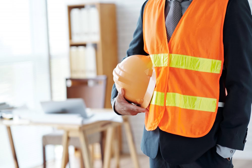 Construction worker in a bright orange safety vest holding a yellow hard hat in an office setting