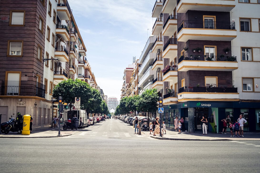 Urban street lined with mid-rise apartment buildings, trees, and people crossing at a crosswalk near a bank on the right.