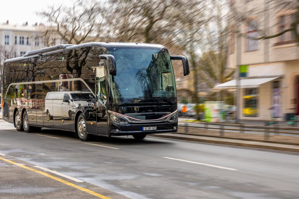 A sleek black tour bus driving down a city street, reflected windows showing surrounding buildings and trees.