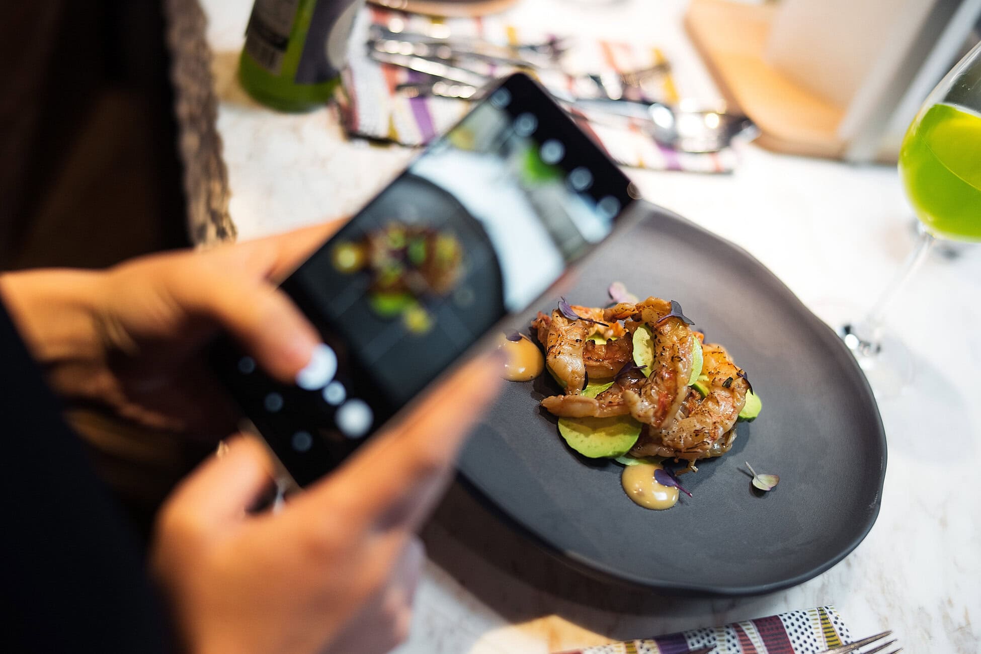 Hand holds a smartphone to take a photo of a plated shrimp dish on a dark plate with lime purée and microgreens beside it.