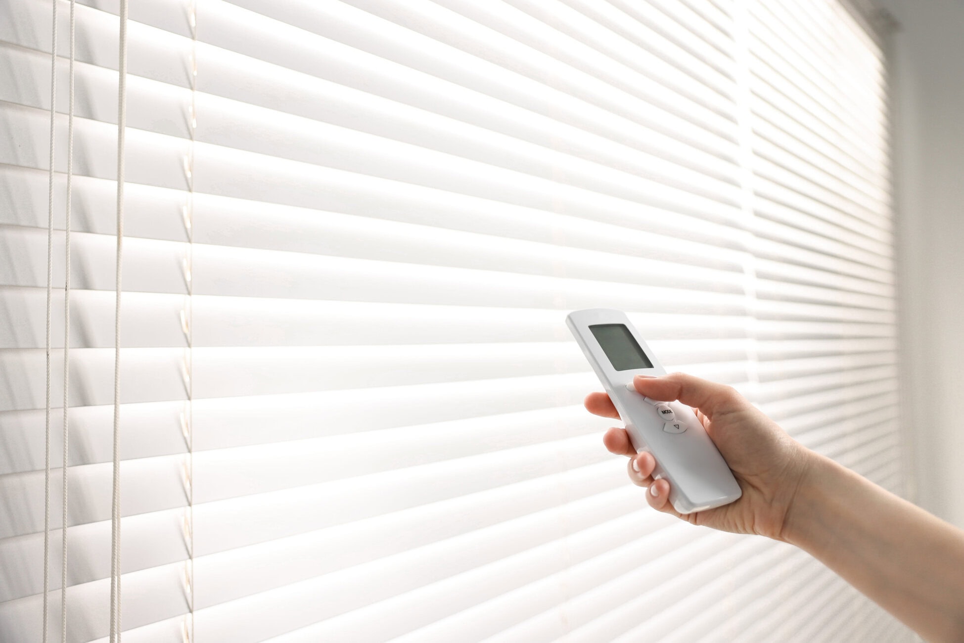 Person holding a white remote control near white horizontal blinds, as if adjusting them