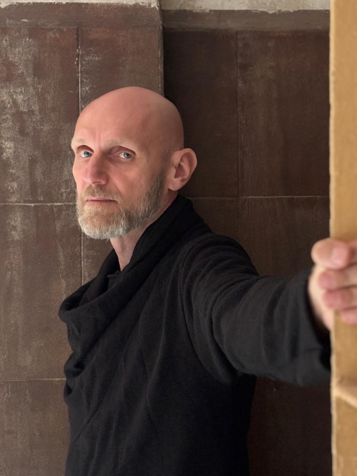 Bald man with a gray beard wearing black clothing, leaning against a brown wall and holding a wooden door frame toward the camera.