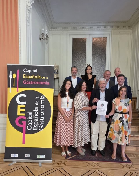 Group of ten people posing indoors beside a banner for the Capital Española de la Gastronomía; one man holds a certificate.