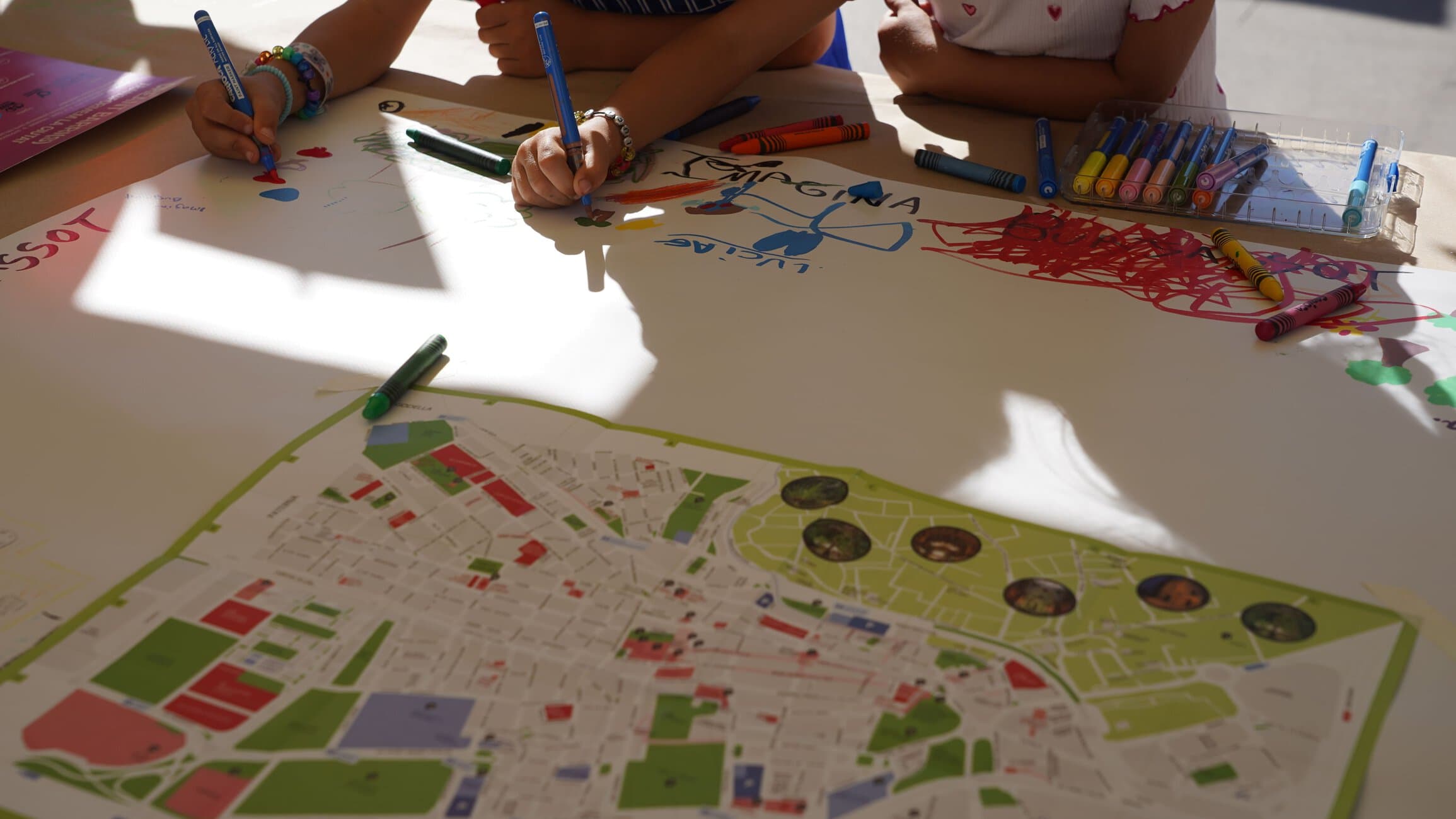Children drawing on a large map poster at a table with blue and green markers, colorful artwork surrounding it.