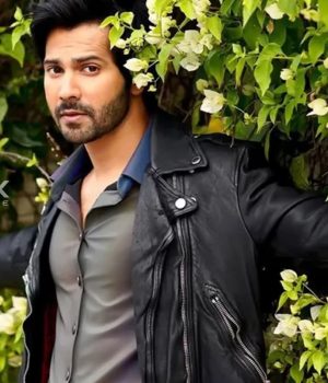 A man with dark hair and a beard wearing a black leather jacket poses among green foliage with white flowers in the background.