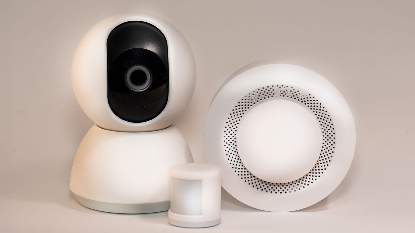 Round white smoke and carbon monoxide alarm beside a smart security camera and sensor on a neutral background