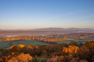 A landscape view of the fall foliage in Northern Dutchess County, with the Kingston-Rhinecliff Bridge and the Catskill Mountains in the distance, from atop the Ferncliff Fire Tower during the golden hour of sunrise on an October morning.