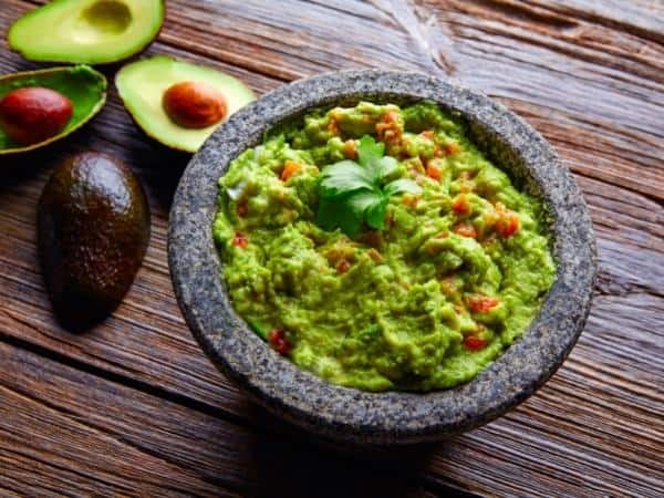 A bowl of guacamole garnished with cilantro sits invitingly amidst halved avocados and a whole one, perfect for those enjoying avocado while following Slimming World on a rustic wooden surface.