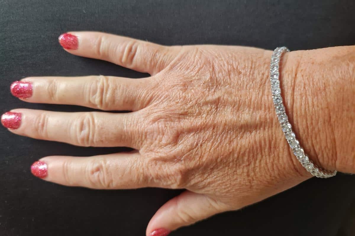 A hand adorned with Carat London jewelry, red glitter nail polish, and a silver bracelet rests elegantly on a black surface.