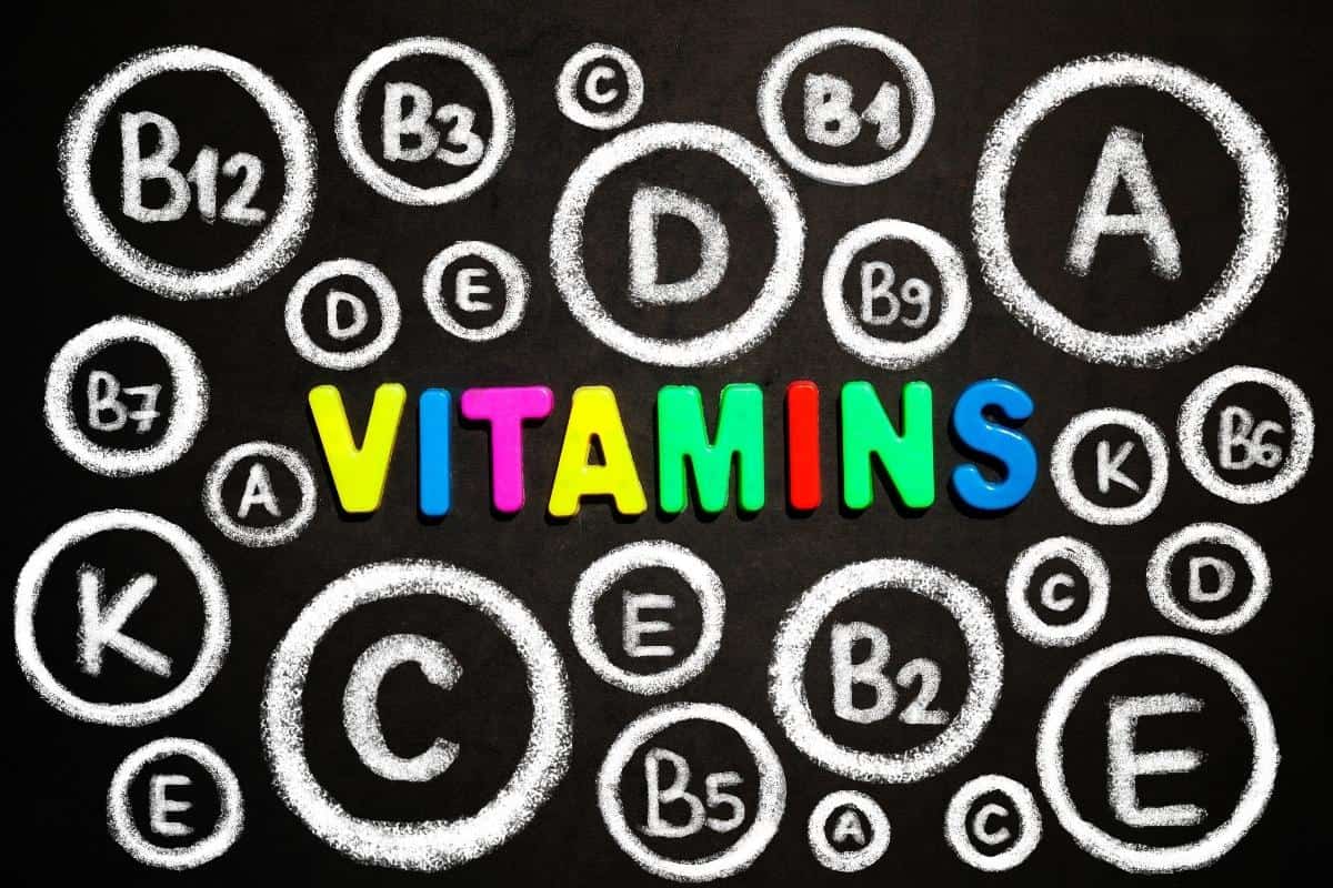 Colorful letters spelling "VITAMINS" are surrounded by chalk-drawn circles featuring different vitamin names and types of vitamin supplements on a black background.