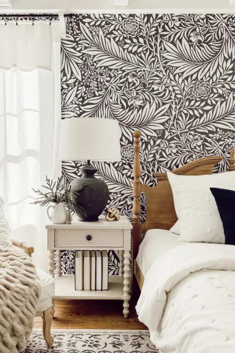 The bedroom features a white bed complemented by a wooden nightstand. Black and white floral wallpaper adds elegance, while a table lamp and decorative plants bring warmth to the space.