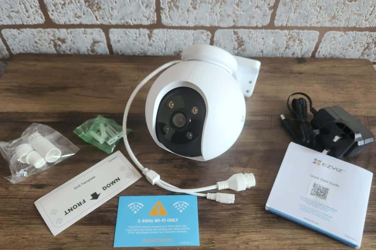 The EZVIZ H8 Pro security camera, complete with cables, mounting hardware, a power adapter, and a quick start guide, is displayed on a wooden surface against a brick wall.