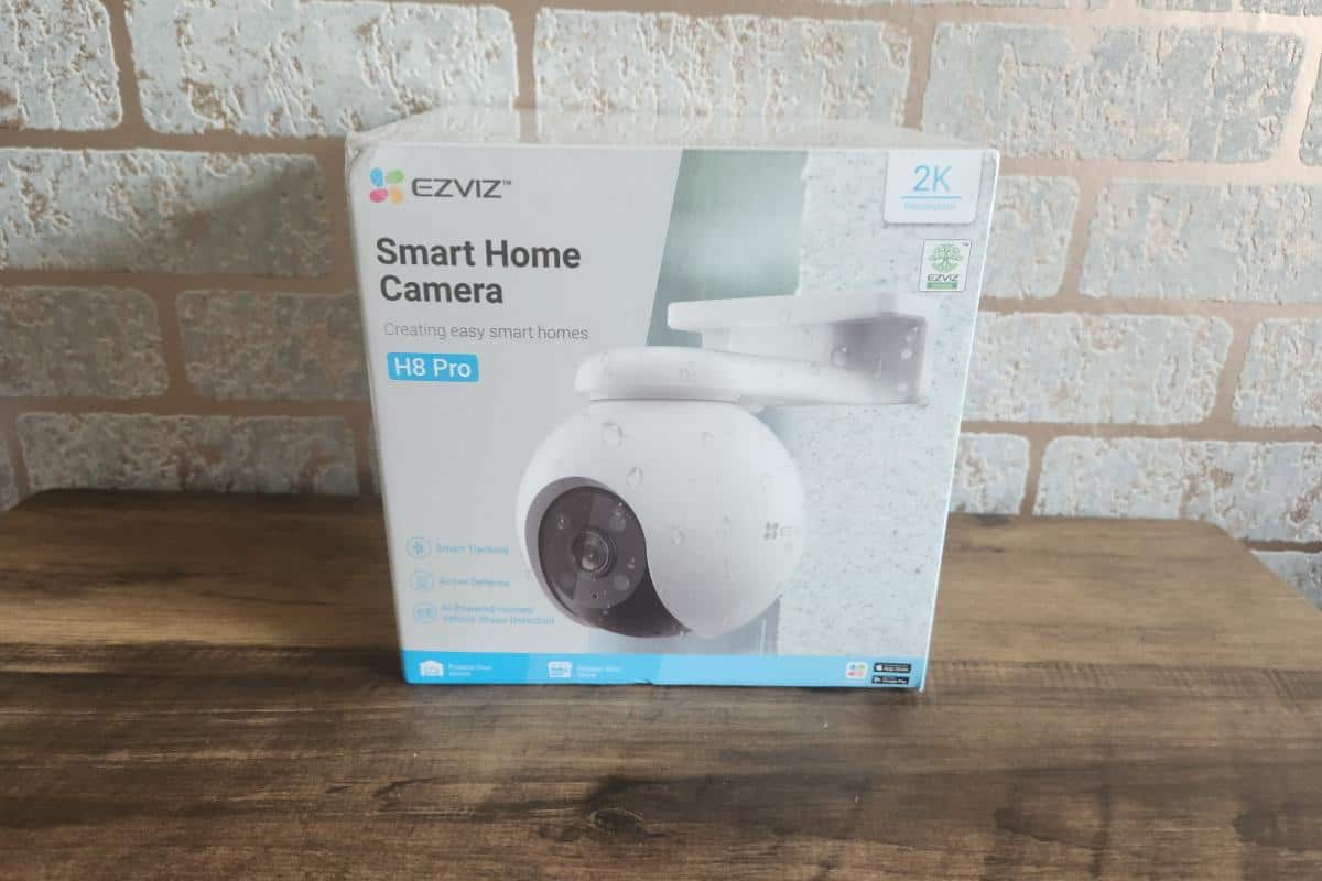 The box of the EZVIZ H8 Pro smart home camera sits elegantly on a wooden surface, framed by a rustic brick wall background.