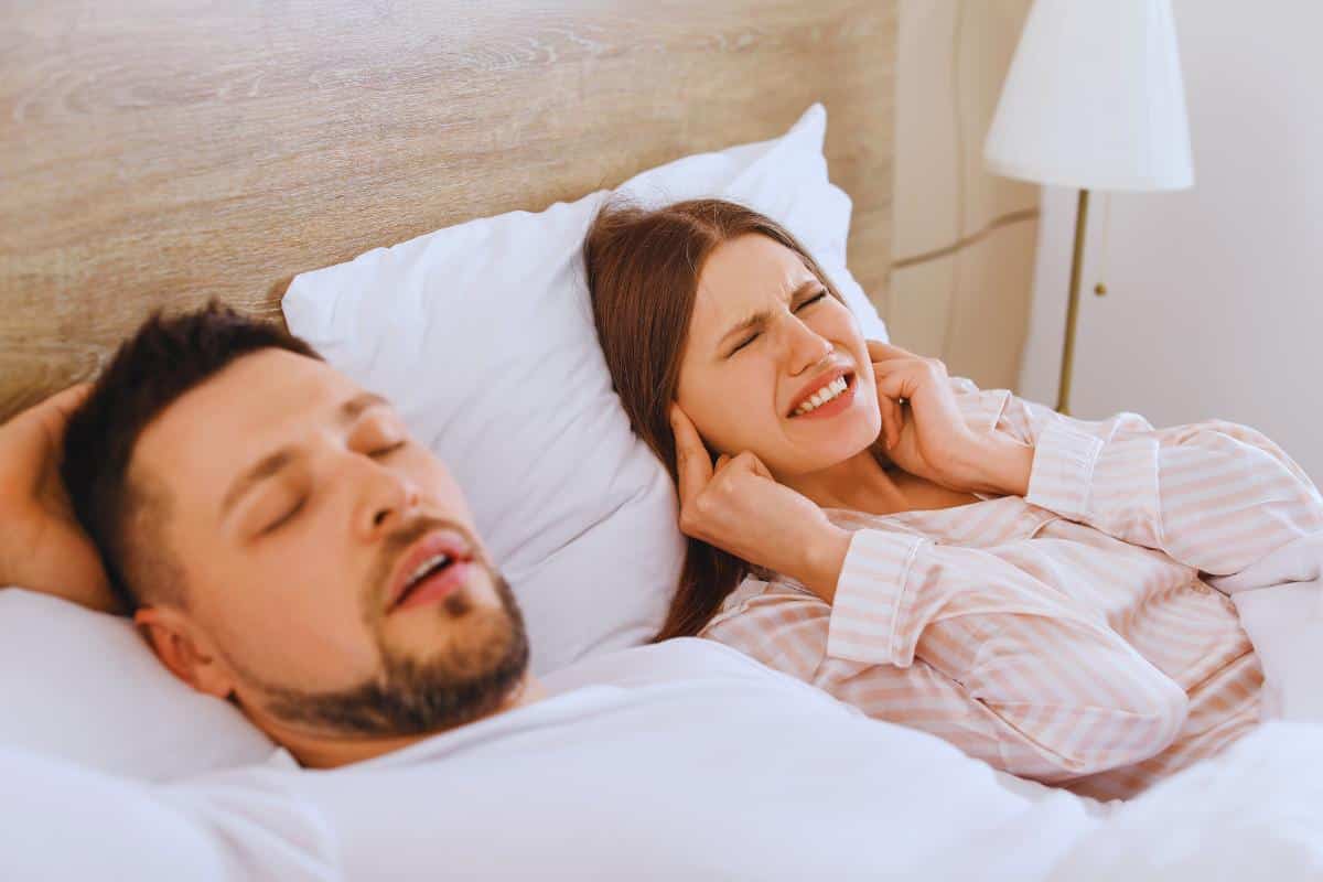 A man is asleep, mouth open, on a bed. Beside him, his partner covers her ears with her fingers, appearing discomforted by his snoring.