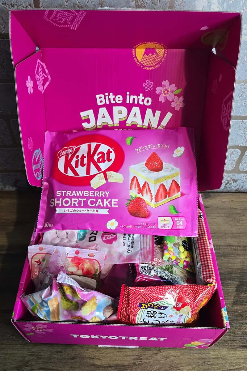 Open pink box with a variety of Japanese snacks, perfect for any gift guide, prominently featuring a KitKat Strawberry Shortcake package. Box lid reads, "Bite into Japan!" An ideal Mother's Day gift to delight mum with flavors from afar.