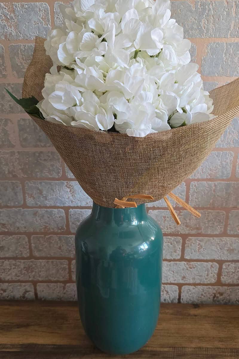 A bouquet of white flowers wrapped in brown paper, thoughtfully placed in a tall, green vase against a light brick wall—perfect for your Mother's Day gift guide.