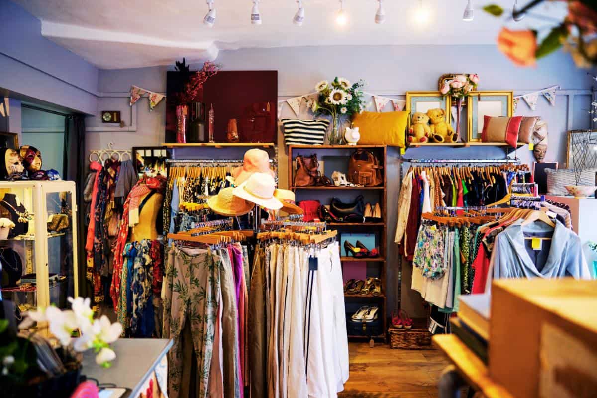 A small boutique interior with racks of colorful clothing, hats, and accessories. Shelves display bags and decorative items. The space is well-lit with spotlights, and floral decor is visible throughout the store.