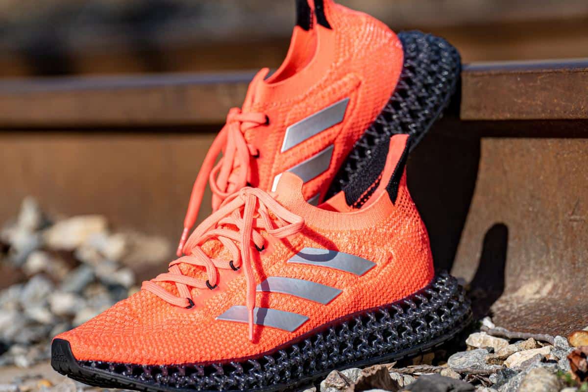 A pair of bright orange Adidas running shoes with silver stripes and black textured soles are placed on gravel beside a metal rail—perfect for adding flair to your autumn workout clothes or any smart woman's guide to fitness style.