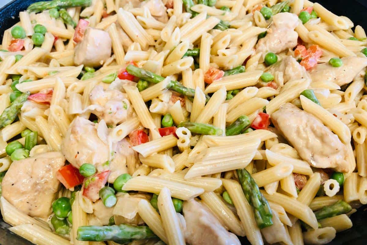 A close-up of creamy, healthy pasta featuring penne mixed with pieces of chicken, green peas, asparagus, and red bell pepper in a skillet.