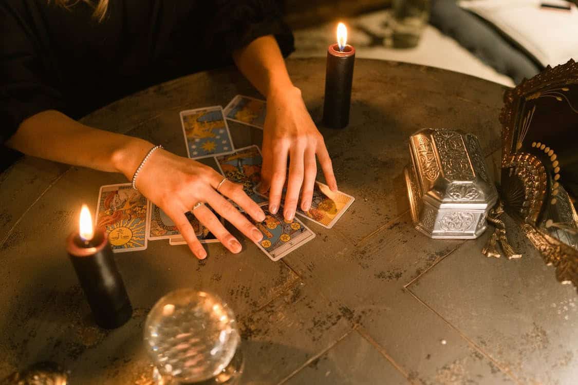 A person arranges tarot cards on a round table lit by two black candles, with a crystal ball and ornate silver box nearby, creating a mystical atmosphere.