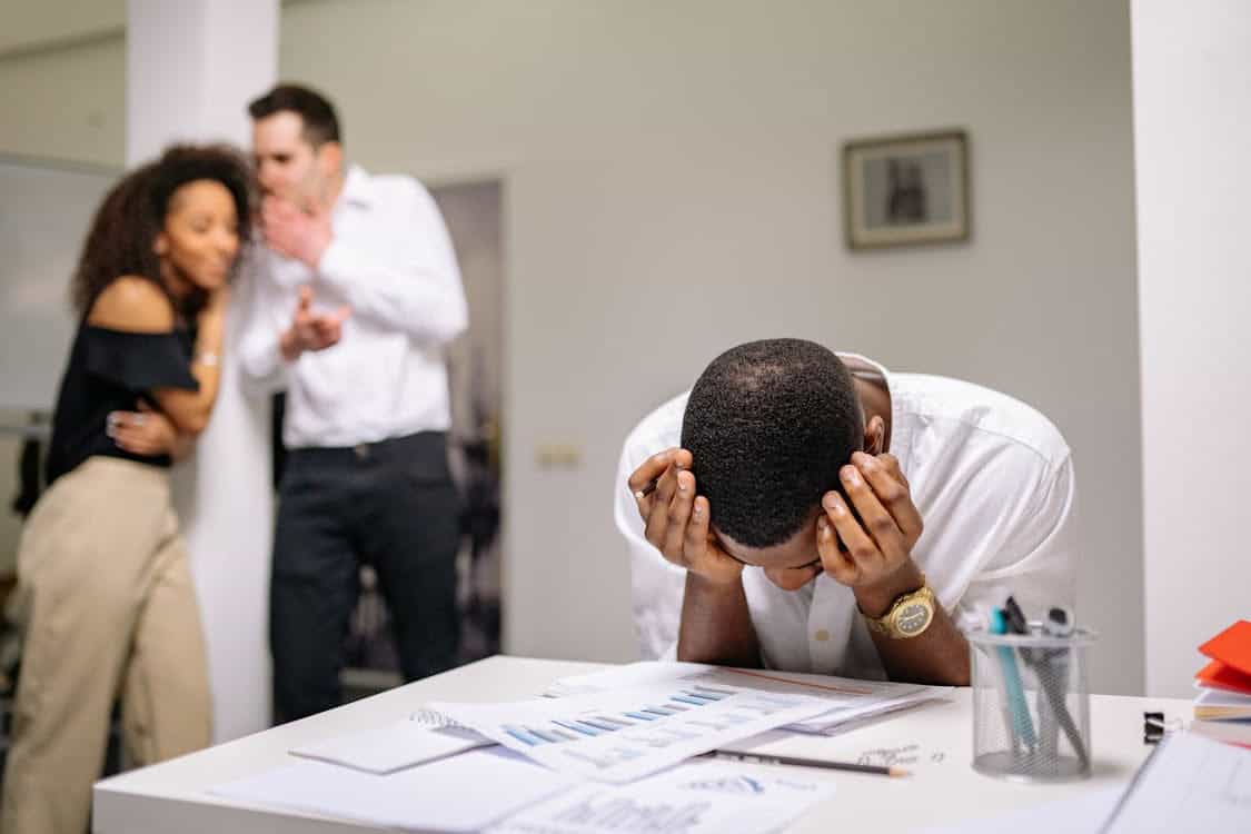 A man sits at a desk with his head in his hands, looking stressed, while two colleagues stand in the background whispering to each other, suggesting gossip or workplace tension.