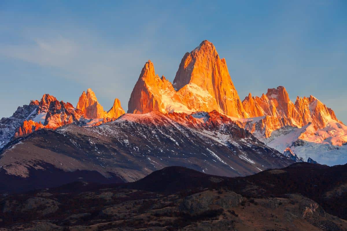 Jagged mountain peaks, topped with snow, glow orange in the warm light of sunrise or sunset. The lower slopes are shadowed and rugged, with patches of forest and rocky land in the foreground.
