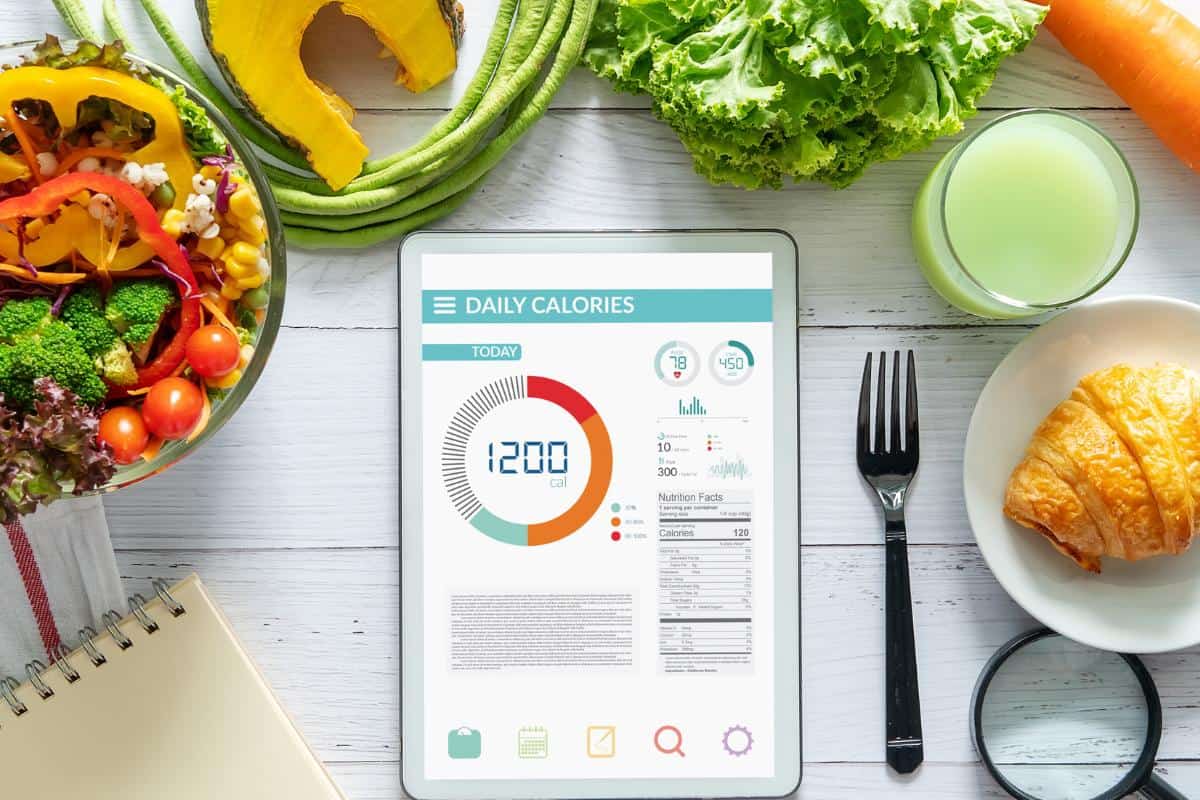 A tablet displaying a daily calorie tracker for healthy weight loss sits on a white table surrounded by vegetables, a croissant, a glass of green juice, a fork, and a notepad with a pen.