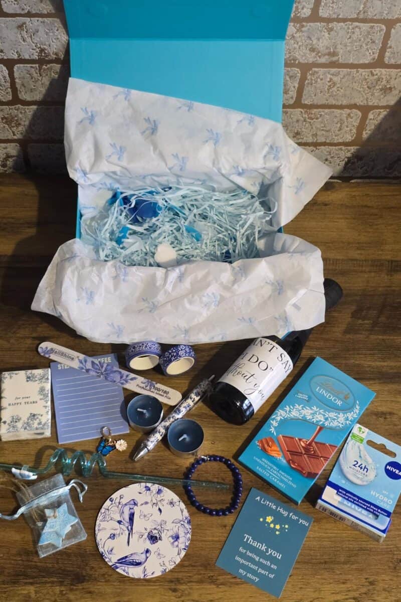A gift box for bridesmaids with blue and white items on a wooden table, including a mug, candle, pen, chocolate, bracelet, Nivea cream, notepad, matches, thank you note, and shredded paper filling—perfect for your “How I Asked” moment.
