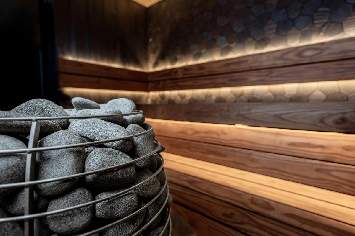 A close-up of sauna stones in a metal basket inside a modern sauna room with wooden benches and warm ambient lighting, highlighting how saunas can support relaxation and weight loss.